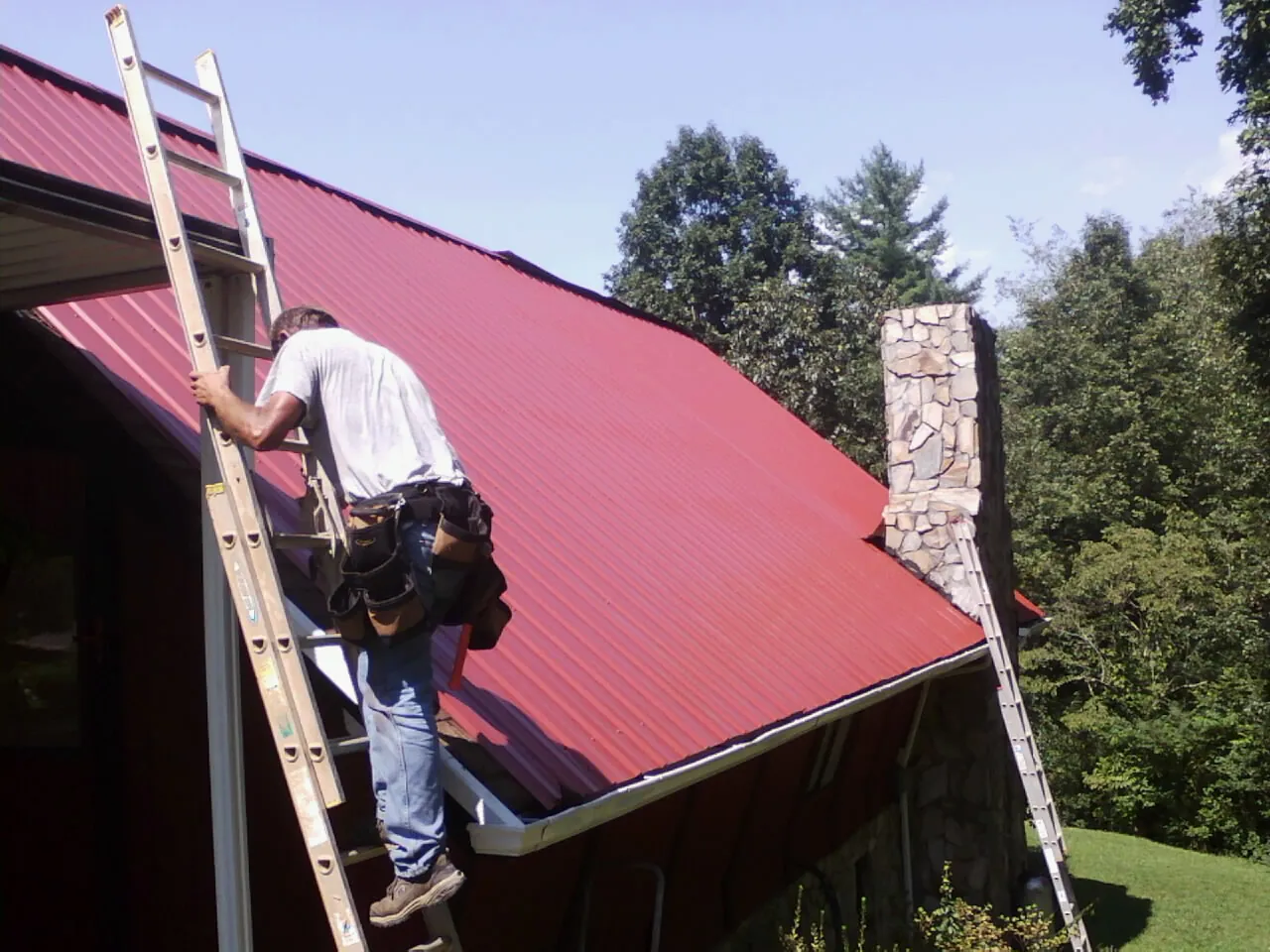 Roofer on a ladder beside a red metal roof and stone chimney — Soffit & Fascia Repair by Roofing Brothers in Westford